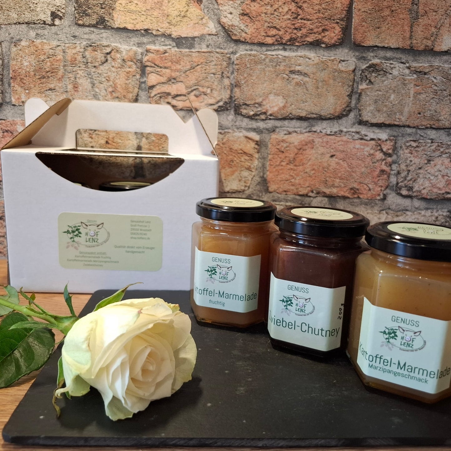 Drei Gläser aus dem Hof Lenz Genusspaket mit Kartoffel-Marmelade fruchtig, Zwiebel-Chutney und Kartoffel-Marmelade Marzipangeschmack, dekoriert mit weißer Rose vor Geschenkbox.