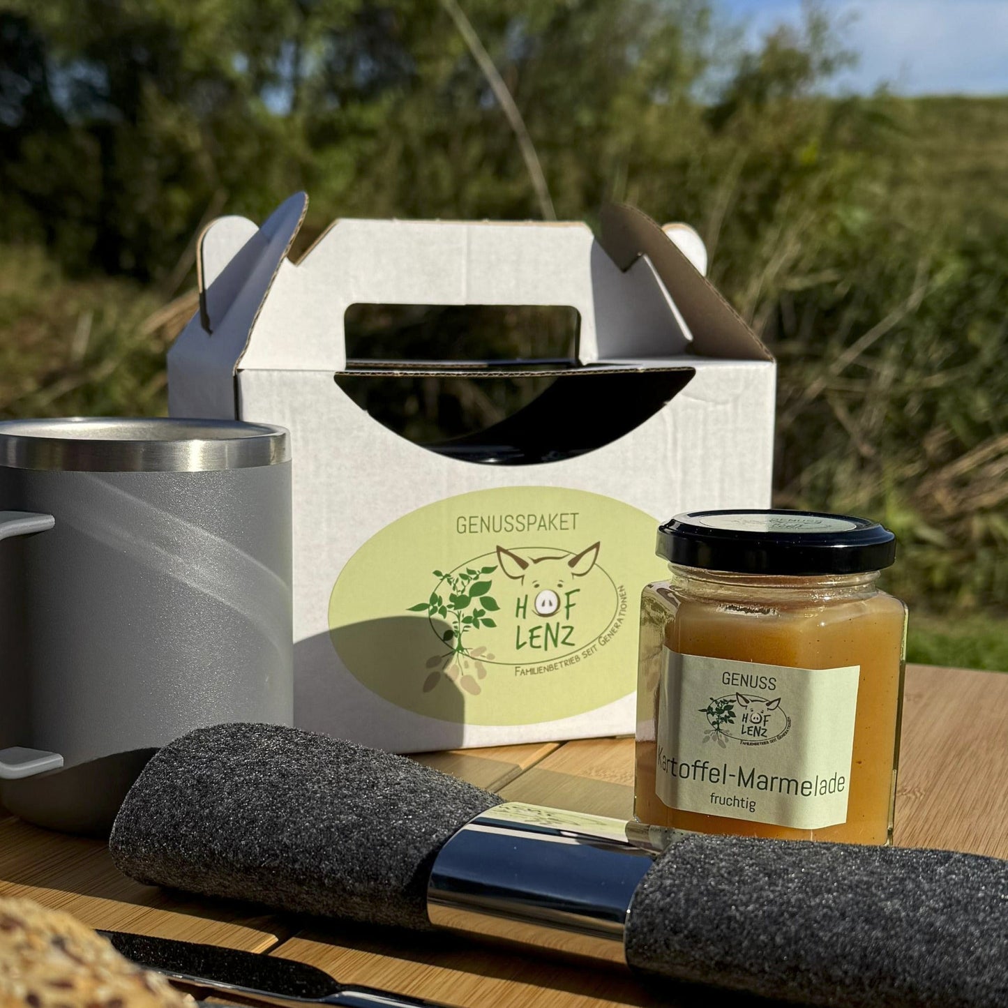 Genusspaket vom Hof Lenz beim Picknick mit Brötchen, Früchten und Getränken auf Holztisch im Grünen, Glas Kartoffel-Marmelade im Vordergrund.